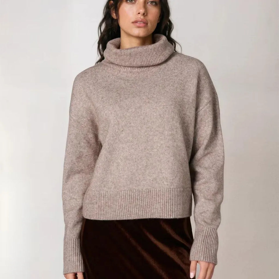 A woman wearing a light taupe knit turtleneck sweater and a dark brown velvet skirt, standing against a plain light gray studio background.