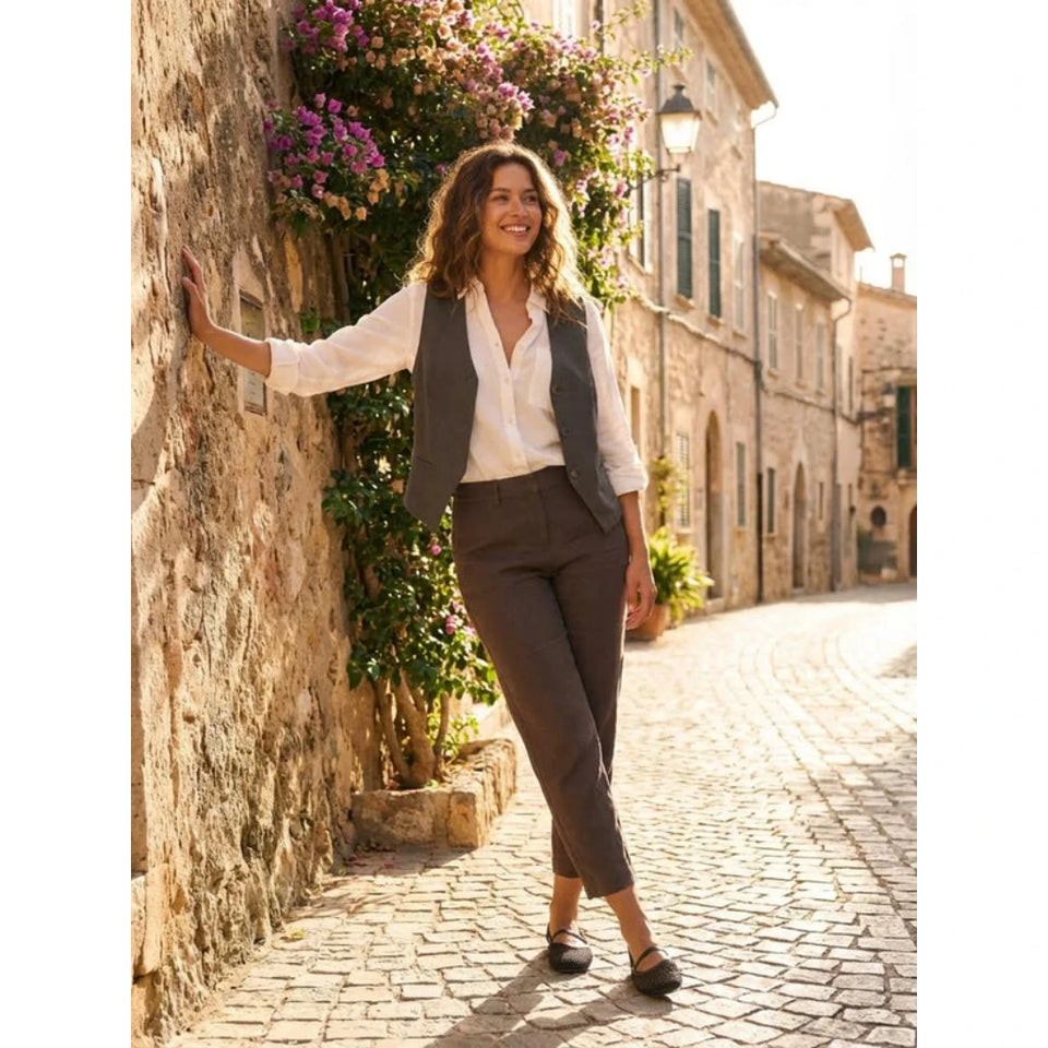 A woman stands on a sunlit cobblestone street wearing a white button-up shirt, a dark charcoal vest, dark brown cropped trousers, and dark loafers, leaning against a rustic stone wall with purple flowers and buildings in the background.