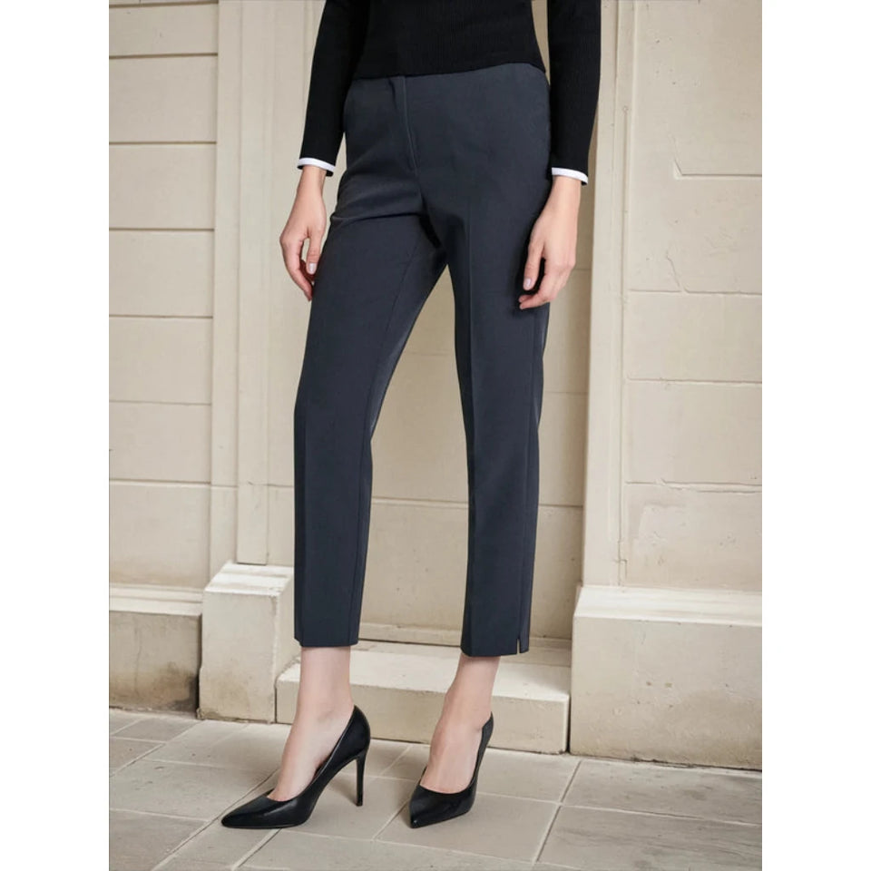 A model wearing dark charcoal gray cropped trousers, a black long-sleeve top with white cuffs, and black pointed-toe heels, standing against a beige stone wall.