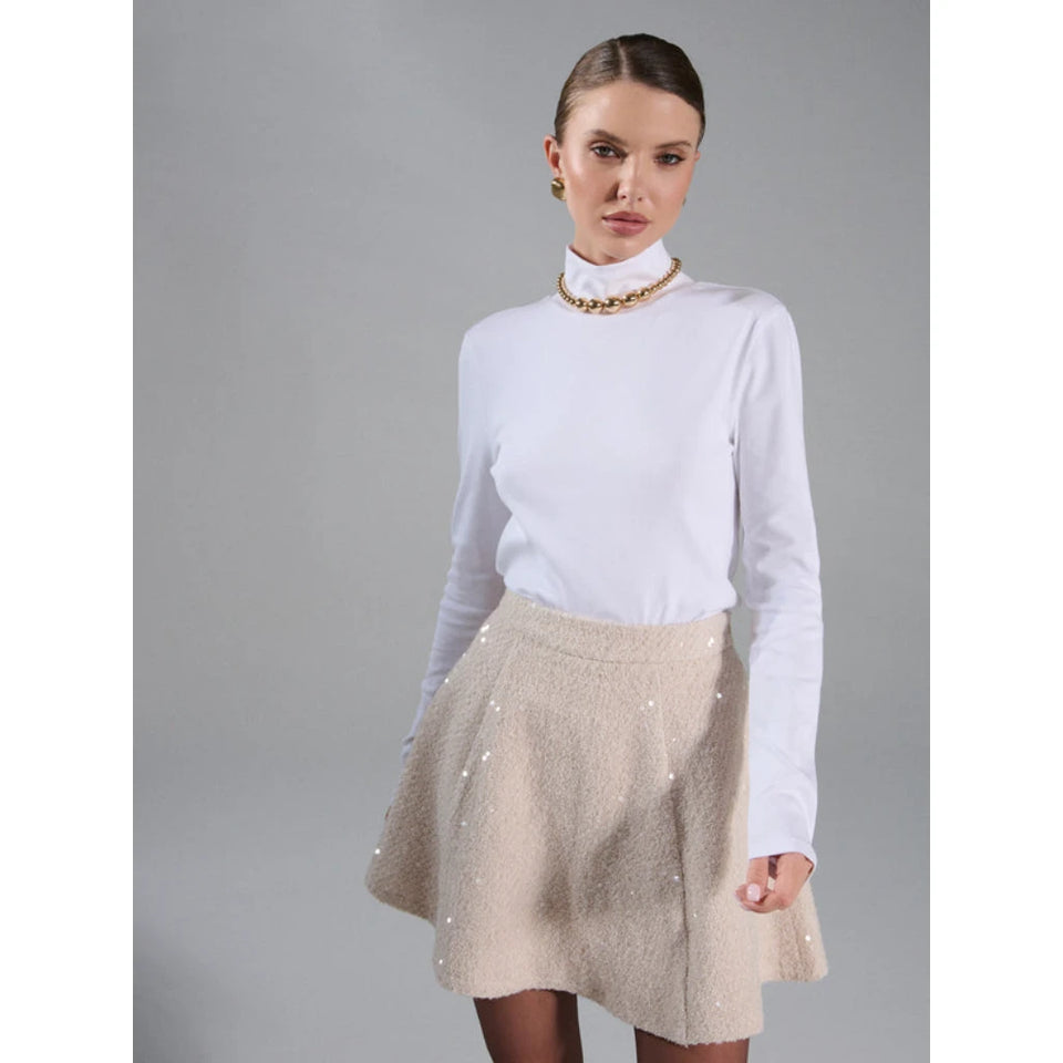 A model wearing a white long-sleeve mock neck top and a beige textured flared mini skirt with sequins, set against a solid gray studio background.