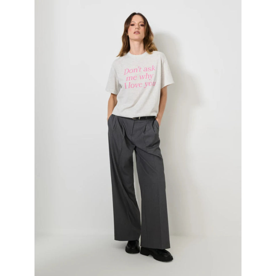 A woman stands against a plain light gray studio wall wearing a light gray graphic T-shirt with pink text reading 'Don't ask me why I love you,' dark gray wide-leg trousers, and black shoes.