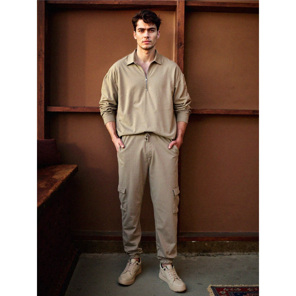 A man modeling a matching beige long-sleeve quarter-zip pullover and cargo pants set, standing in front of a brown wooden wall.