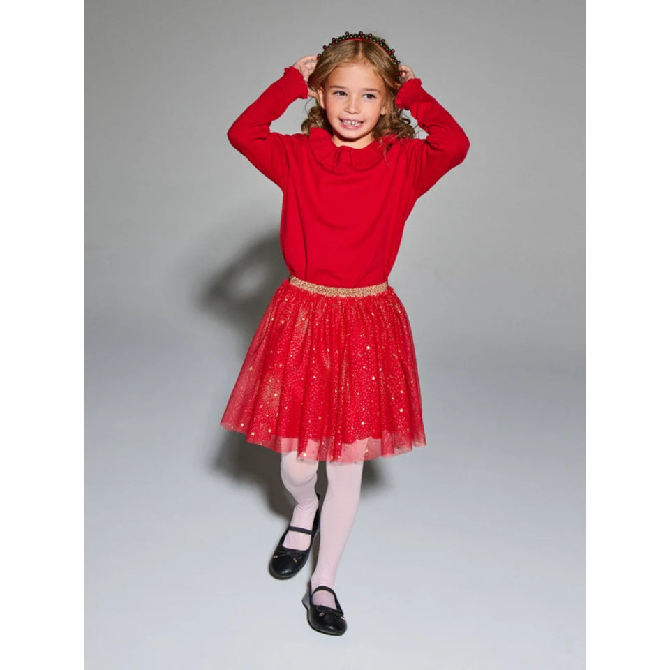 A young girl modeling a festive red holiday outfit consisting of a long-sleeve cowl-neck knit top and a sparkly tulle tutu skirt with a gold glittery waistband, paired with white tights and black shoes, against a neutral gray studio background.
