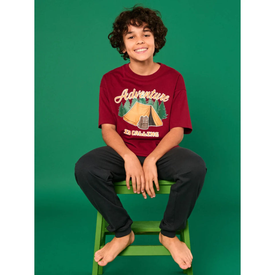 A smiling boy with curly dark hair sits barefoot on a green stool against a solid green studio background, wearing a burgundy graphic T-shirt with 'Adventure 20 CALL2026' and a tent illustration, paired with dark pants.