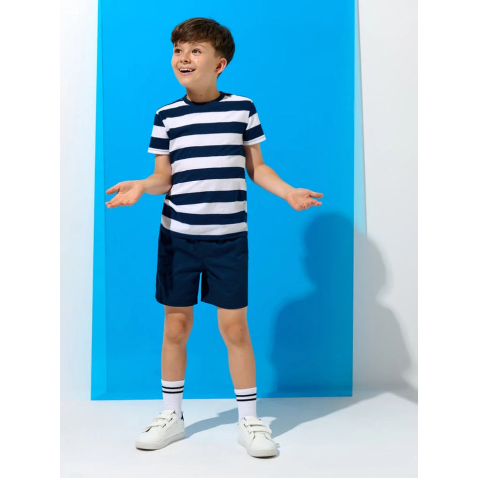 A young boy modeling a navy blue and white striped short-sleeve t-shirt with matching navy shorts, white socks, and white sneakers, standing against a bright blue and white studio backdrop.