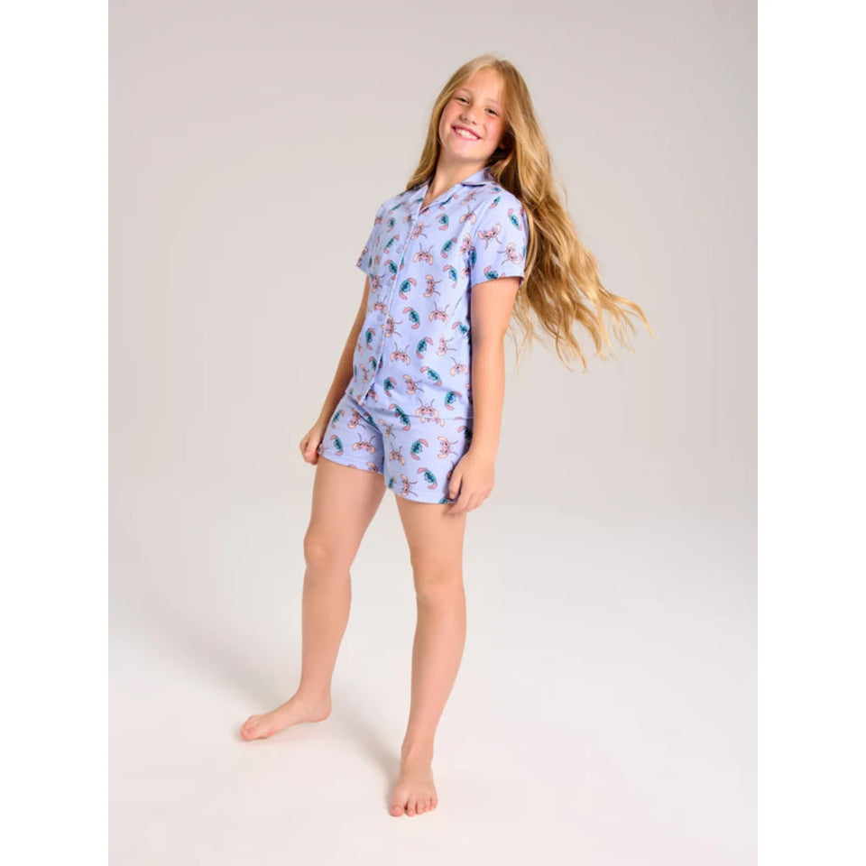 A young girl with long blonde hair stands barefoot against a light gray studio background, wearing a short-sleeve collared pajama top and matching shorts in light blue with a pink and multicolor tropical print.