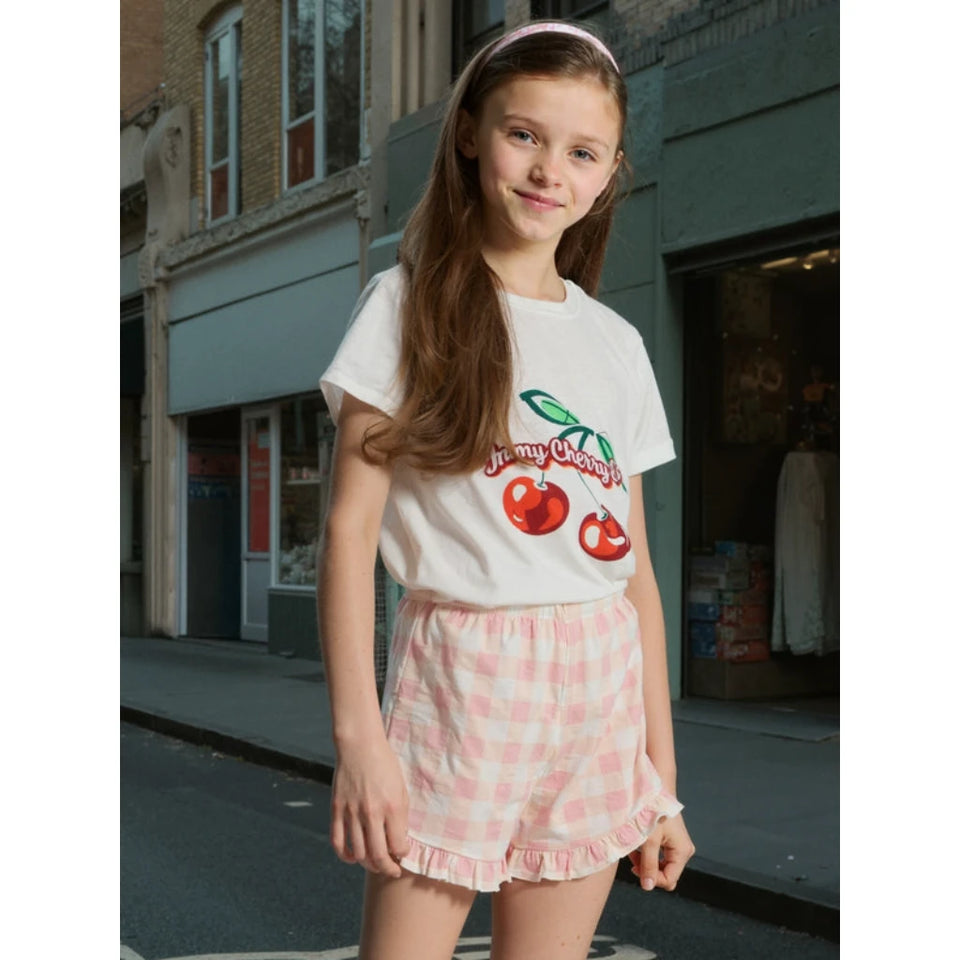 A young girl stands on a city street wearing a white graphic T-shirt with a red cherry design and the words 'Jenny Cherry,' paired with pink and white gingham ruffled shorts.