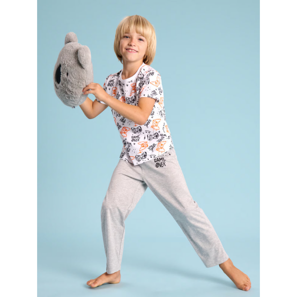A young boy wearing a matching pajama set consisting of a white short-sleeve t-shirt with a colorful cartoon animal print and light gray sweatpants, posing dynamically while holding a gray plush toy against a solid light blue background.