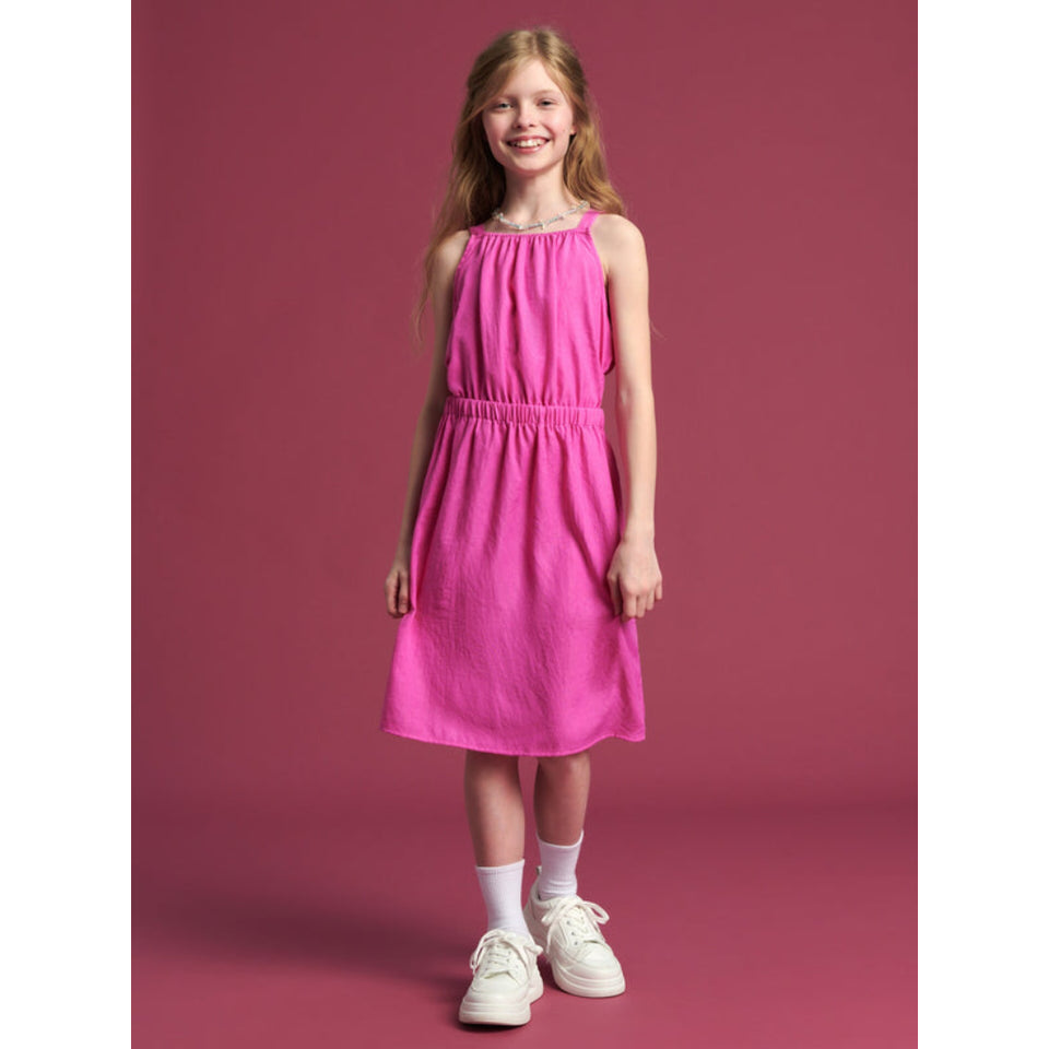 A young girl with long light brown hair stands against a solid dusty rose pink studio background, wearing a sleeveless bright pink midi dress, white socks, and white sneakers.