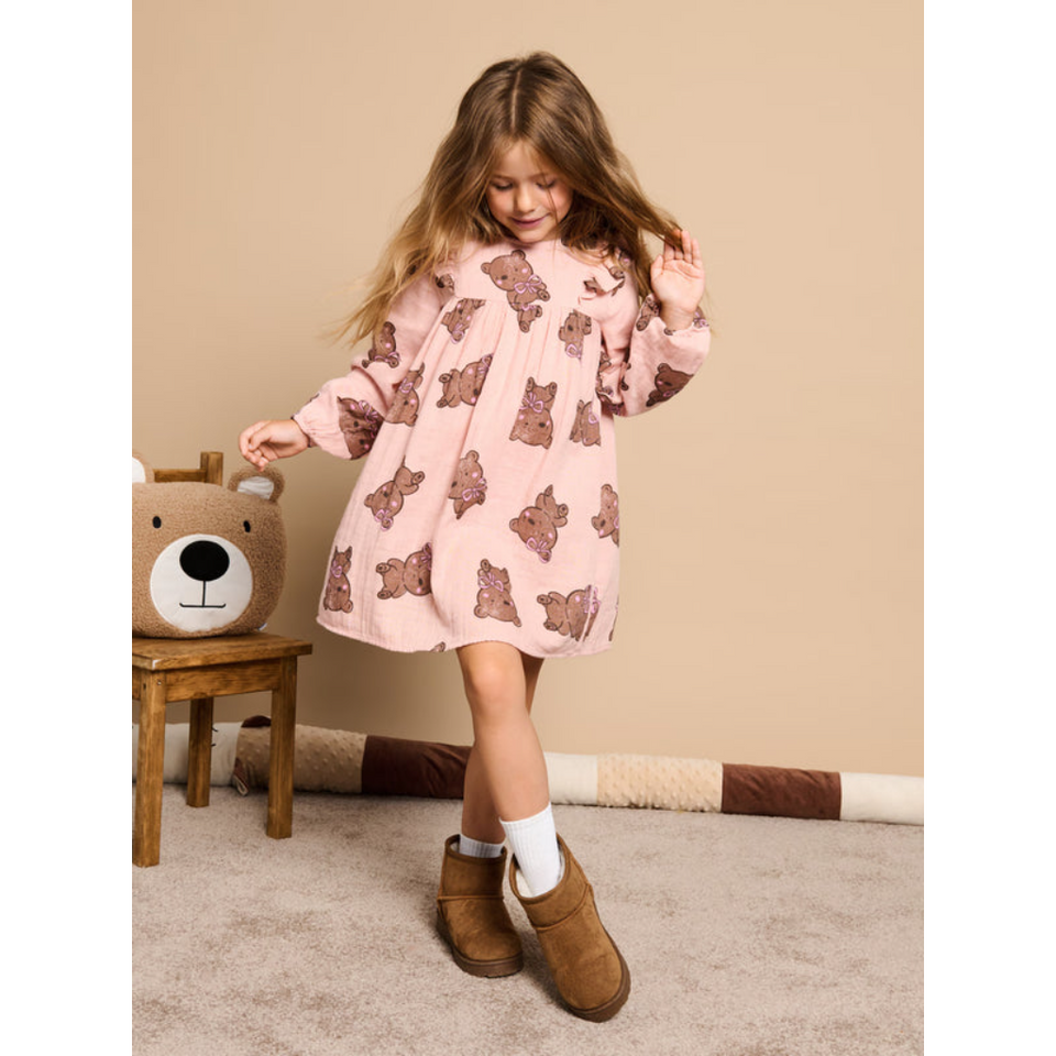 A young girl stands in a room wearing a light pink long-sleeve dress with a brown teddy bear print, white socks, and brown suede boots. A small wooden stool with a plush bear head sits to her left against a beige wall.
