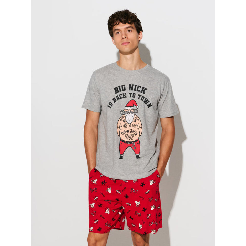 A man modeling a heather gray graphic t-shirt and red patterned shorts against a light gray studio background. The t-shirt features a cartoon graphic of a tattooed man wearing a Santa hat with the text 'BIG NICK IS BACK TO TOWN'.