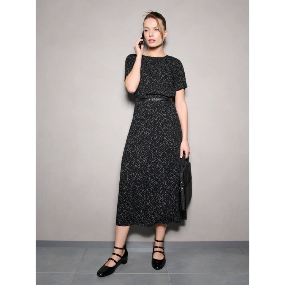 A woman stands against a plain light gray wall wearing a short-sleeve black midi dress with a small white allover print, a thin black belt at the waist, and black Mary Jane-style shoes. She is holding a black handbag and appears to be talking on a phone.