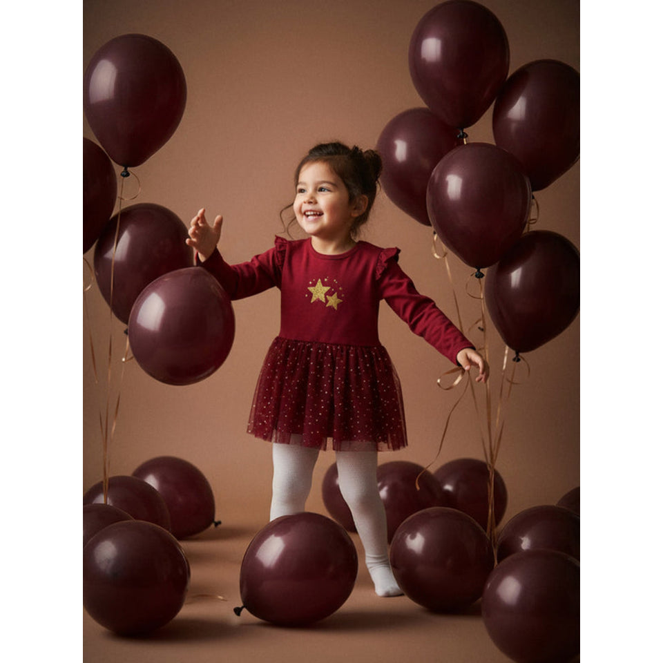 A young girl stands smiling among dark burgundy balloons, wearing a long-sleeve burgundy dress with a gold star on the chest, a sparkly tulle skirt, white leggings, and white socks against a warm brown studio backdrop.