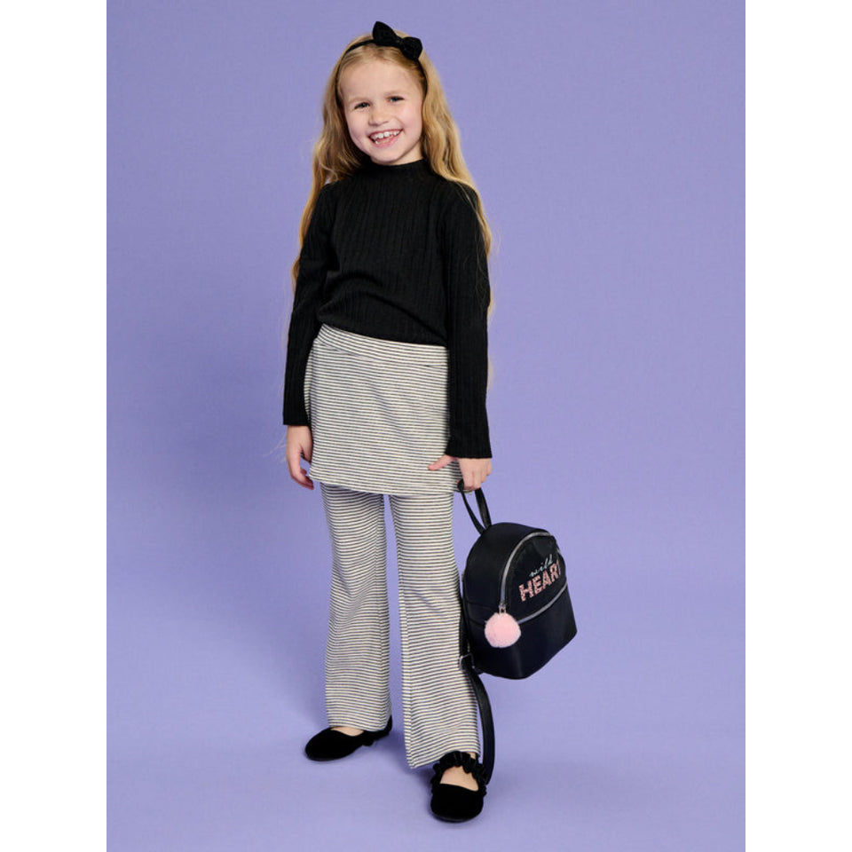 A young girl stands against a solid lavender studio background wearing a black ribbed mock-neck sweater, a gray and white striped mini skirt, and matching gray striped wide-leg pants. She is holding a small black backpack with pink pom-poms and 'HEART' printed on the front.