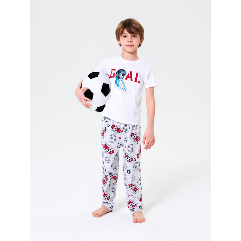 A young boy stands barefoot against a light gray studio background, holding a black-and-white soccer ball. He is wearing a white short-sleeve T-shirt with a red and blue 'GOAL' graphic and white pajama pants covered in a soccer-themed print.