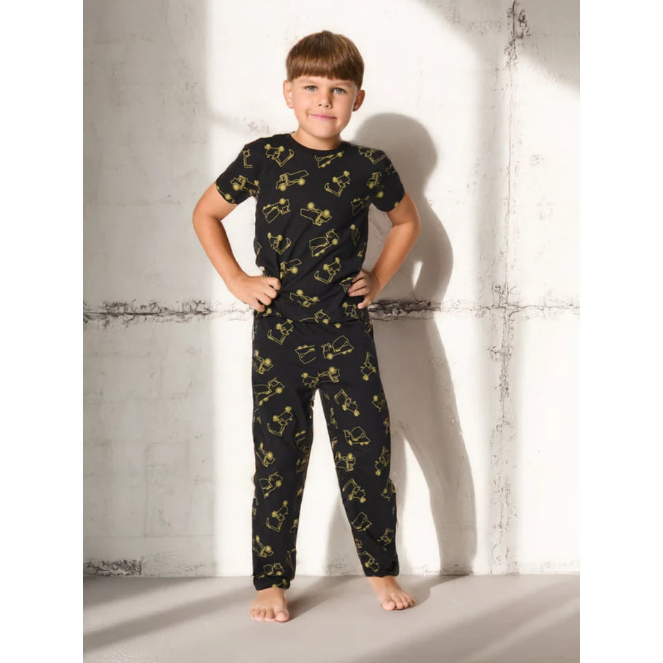 A young boy stands barefoot against a textured concrete wall, wearing a black short-sleeve pajama set with a yellow graphic pattern of cartoon characters.