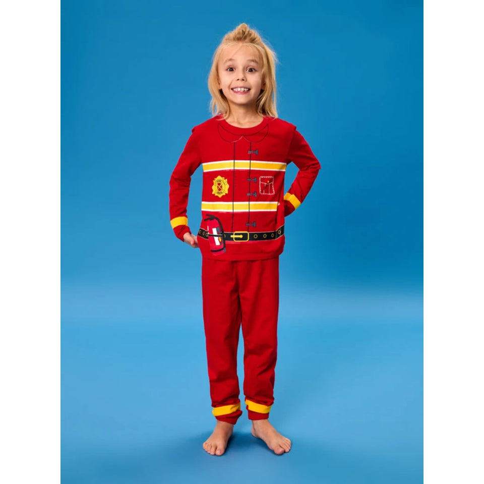 A young girl stands barefoot against a solid blue studio background, wearing a red two-piece pajama set styled like a firefighter uniform with yellow and white stripes, a yellow helmet graphic, and a black belt detail.