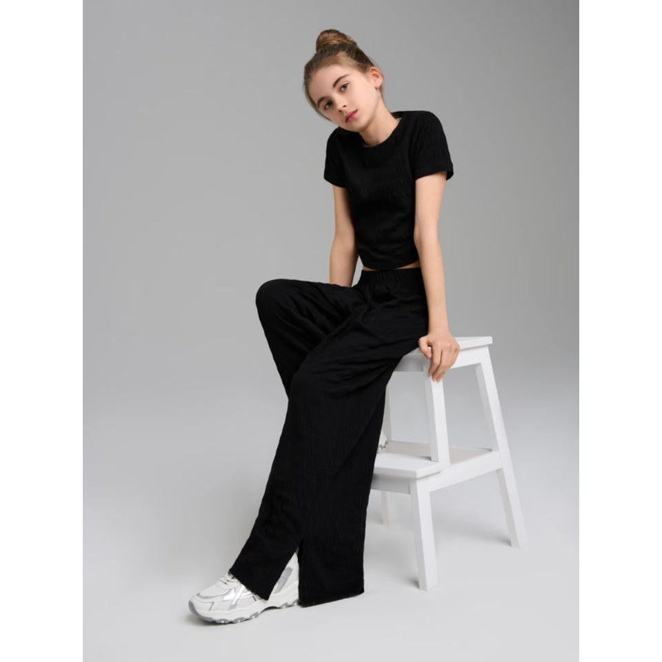 A woman wearing a black short-sleeve crop top and black wide-leg trousers sits on a white stool against a light gray studio background. She is wearing white sneakers.