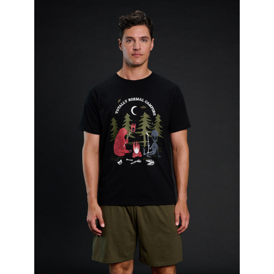 A man modeling a black crewneck t-shirt with a colorful graphic of a campfire scene and the text 'FOOLishly NORMAL CAMPING'. He is wearing olive green shorts against a dark studio background.