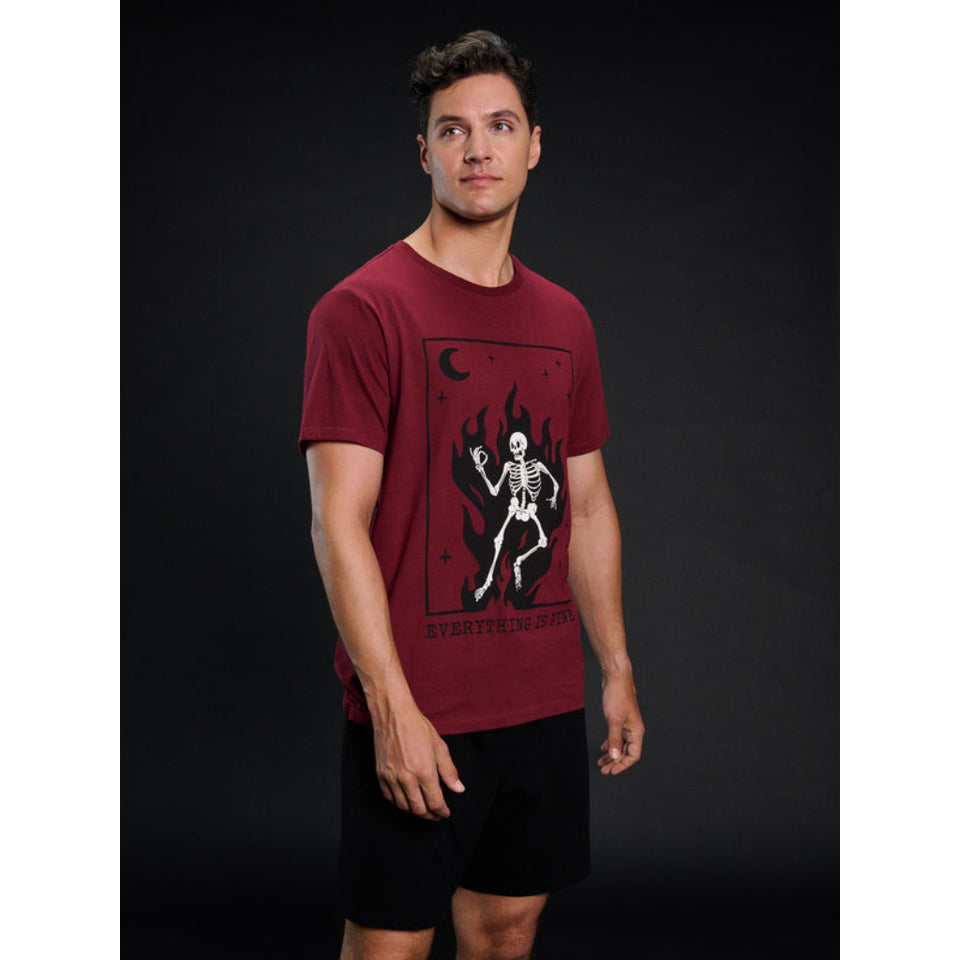 A man modeling a dark red graphic t-shirt featuring a skeleton and flames on the front, paired with black shorts, set against a dark studio background.
