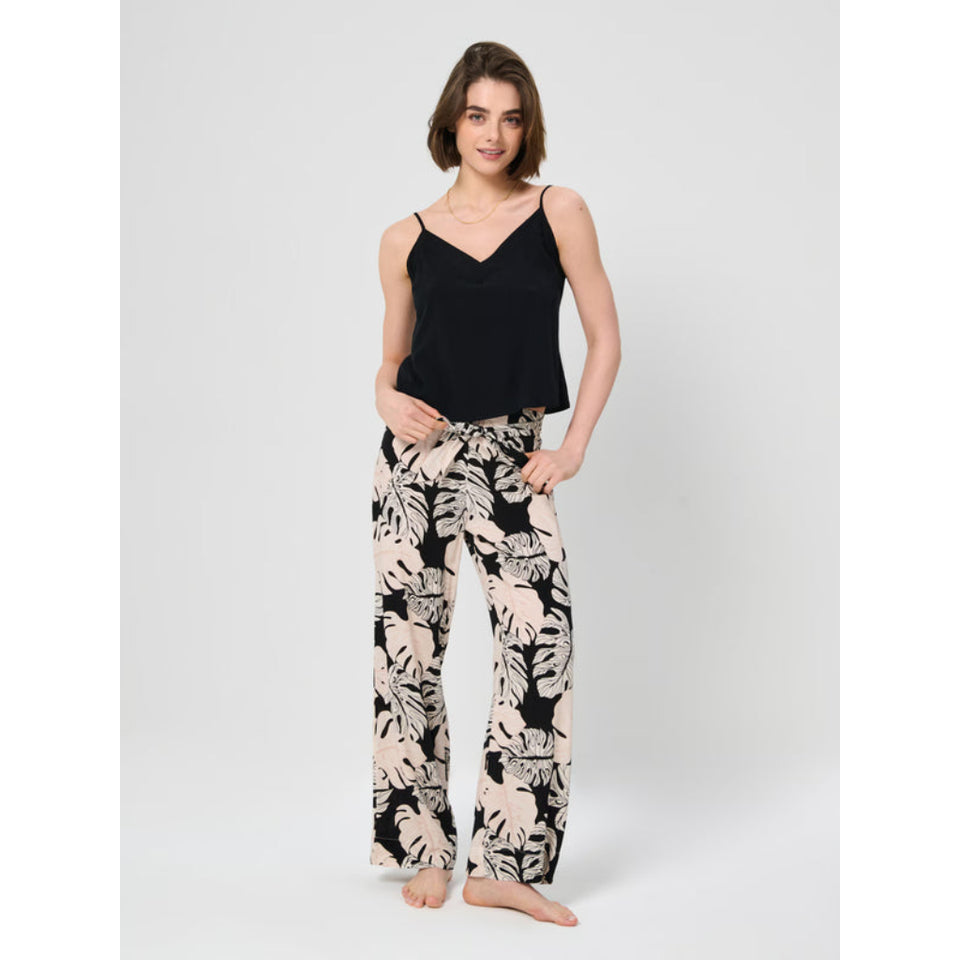A woman modeling a black camisole top and black wide-leg pants with a beige palm leaf print, standing against a light gray studio background.