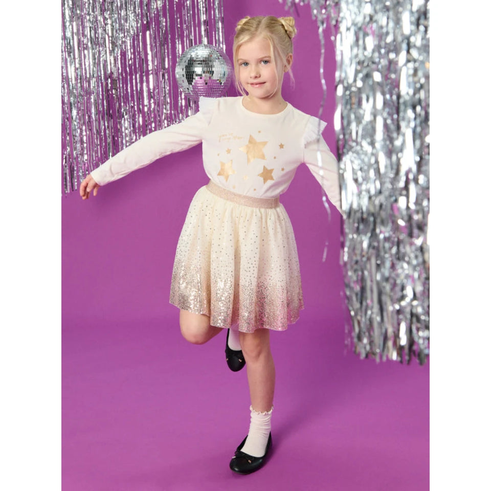 A young girl posing in a white long-sleeve top with gold star graphics and a sparkly champagne-gold sequined skirt, set against a purple background with silver tinsel and a disco ball.
