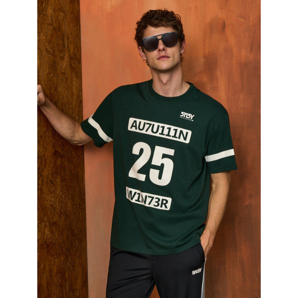 A man wearing a dark green short-sleeve t-shirt with white graphic text and numbers, paired with black pants, posing against a textured orange-brown wooden wall.
