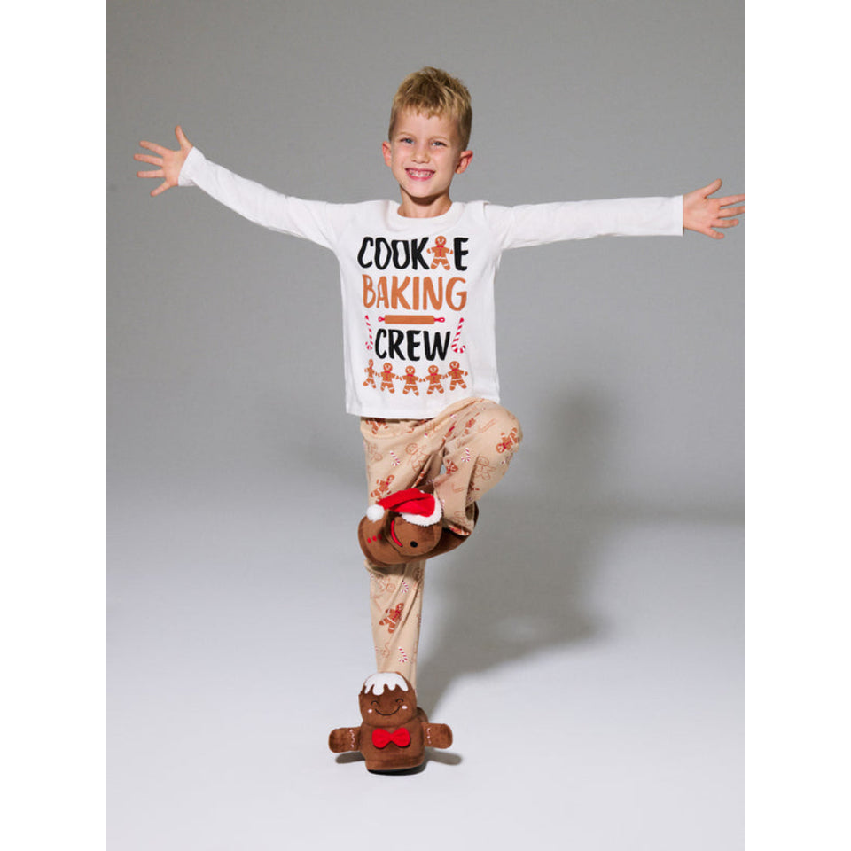 A young boy posing in a white long-sleeve t-shirt with a 'Cookie Baking Crew' graphic and beige patterned pajama pants, balancing on one foot while wearing gingerbread-themed slippers against a gray studio background.