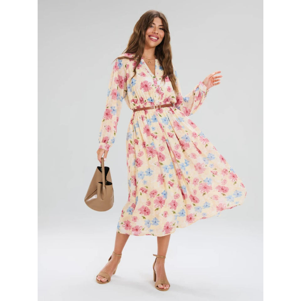 A woman modeling a long-sleeve midi dress with a pink, blue, and yellow floral print on a cream background, featuring a brown belt at the waist. She is holding a brown handbag and wearing beige strappy heels against a light gray studio background.