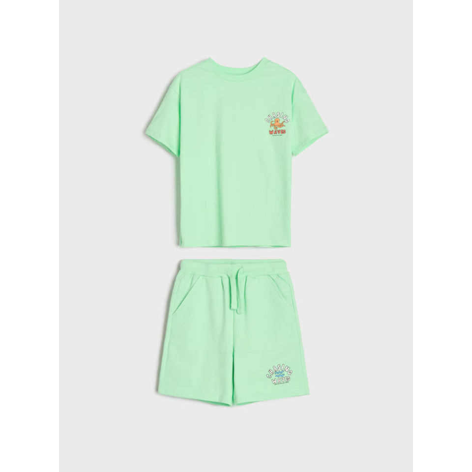 A mint green two-piece lounge set consisting of a short-sleeve t-shirt and matching drawstring shorts, laid flat on a light gray background. The t-shirt features a small colorful graphic on the left chest, and the shorts feature a matching graphic on the lower left leg.