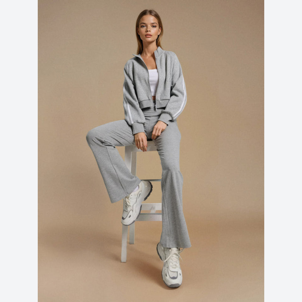 A woman models a light gray two-piece tracksuit consisting of a cropped zip-up jacket with white side stripes and matching wide-leg pants, sitting on a white stool against a solid beige studio background.
