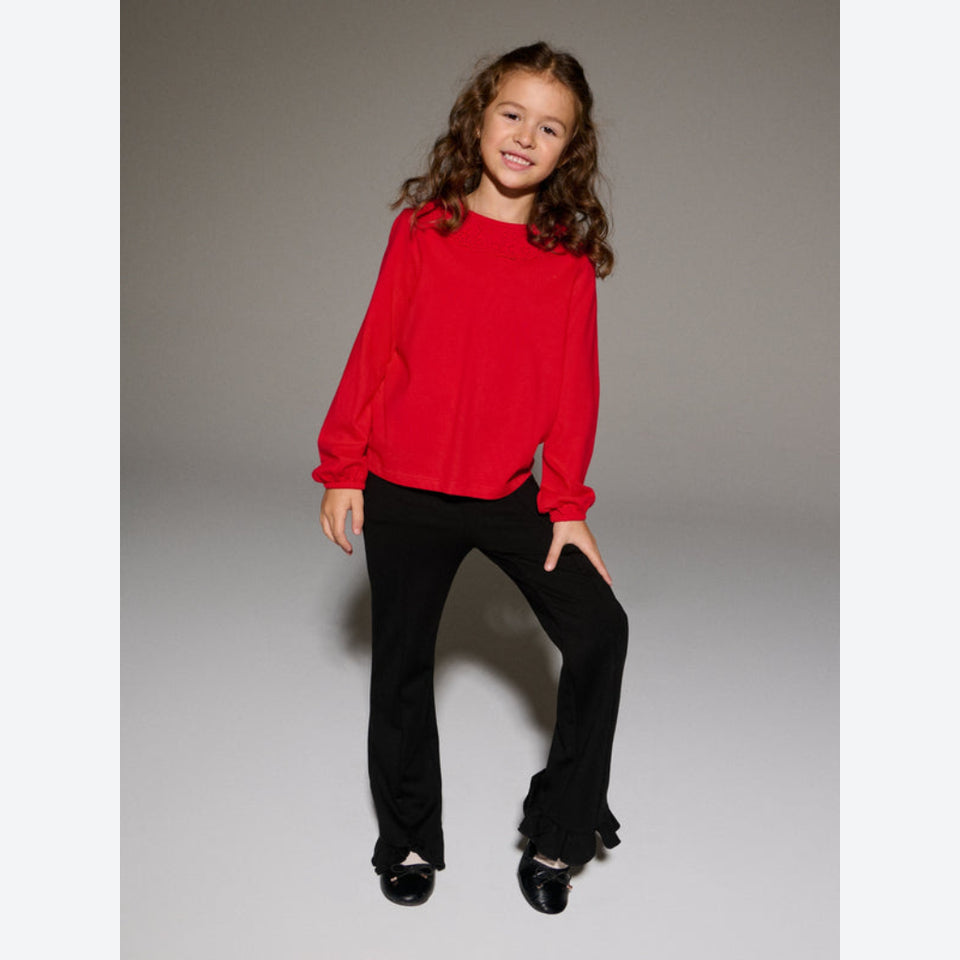 A young girl with long brown hair stands against a neutral gray studio background, wearing a bright red long-sleeve top and black flared pants with ruffled hems.