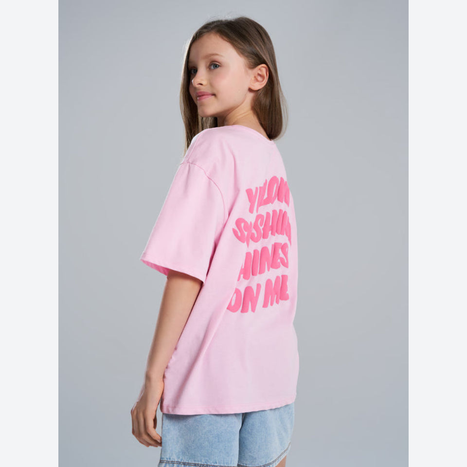 A young girl with brown hair is shown from the back wearing a light pink oversized t-shirt with bright pink text reading 'YLOONG SHIWI AMES ON ME' on the back, paired with light blue denim shorts, against a solid gray studio background.