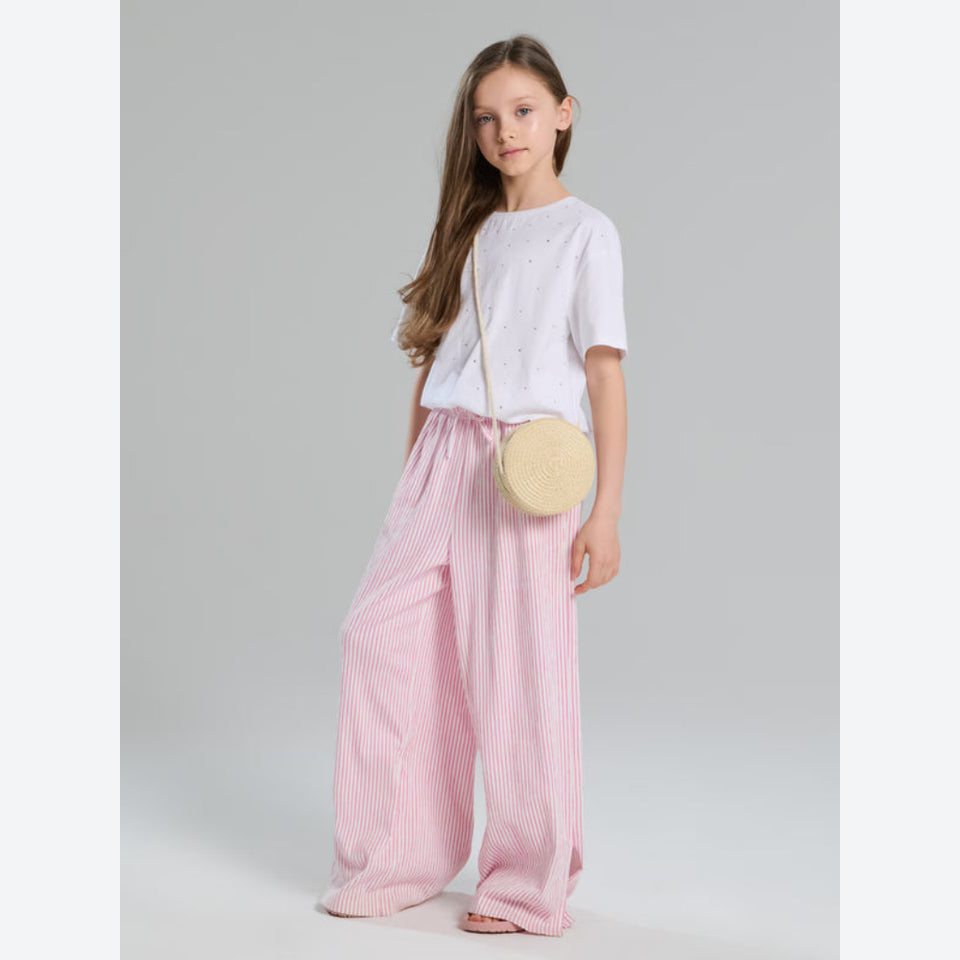 A young girl stands against a light gray studio background wearing a white short-sleeve top with small dot details, pink and white vertically striped wide-leg pants, and a round beige woven crossbody bag.