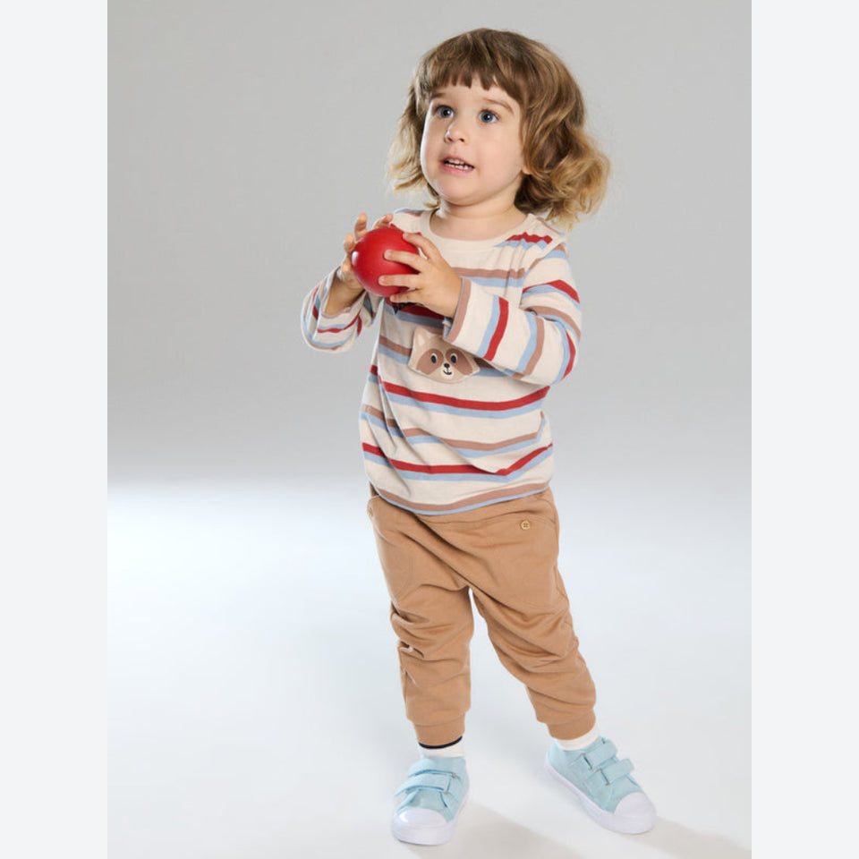 A toddler standing against a light gray studio background, holding a red ball. The child is wearing a cream long-sleeve shirt with thin red, blue, and gray horizontal stripes, a small bear face graphic on the chest, tan pants, and light blue sneakers.
