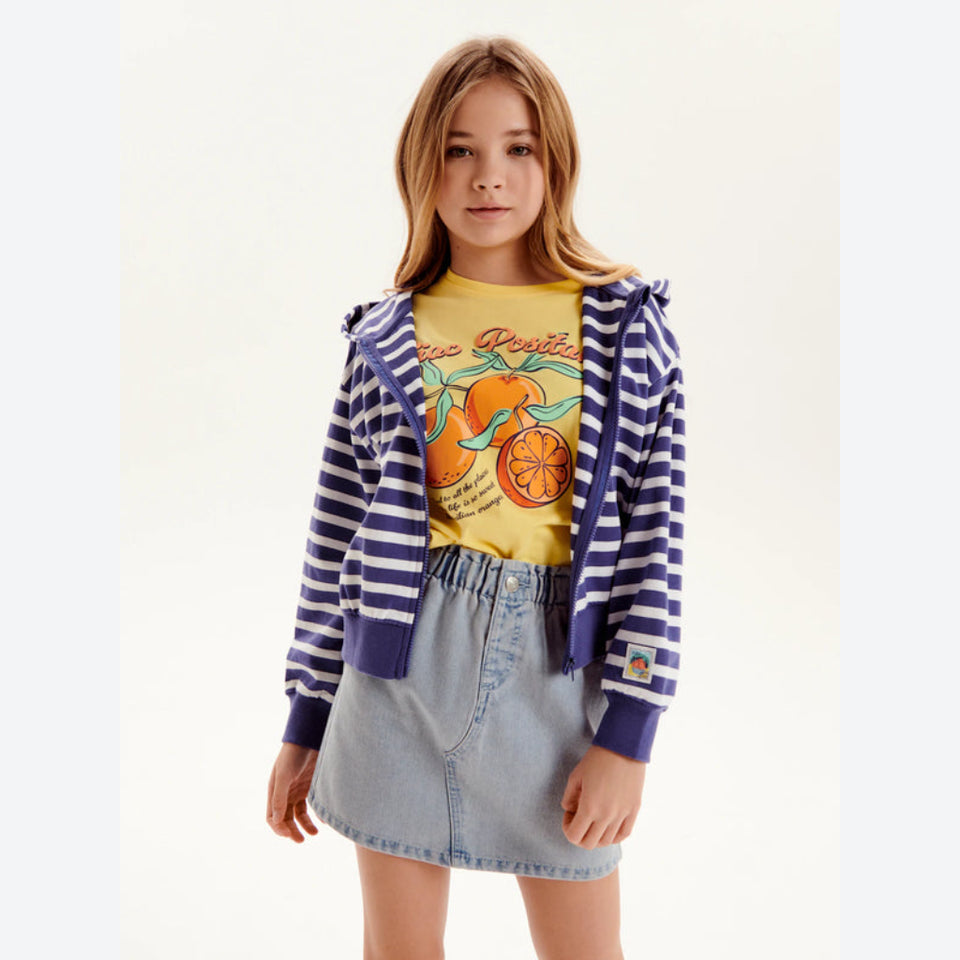 A young girl modeling a striped navy and white zip-up hoodie over a yellow graphic t-shirt and a light blue denim mini skirt, set against a plain off-white studio background.