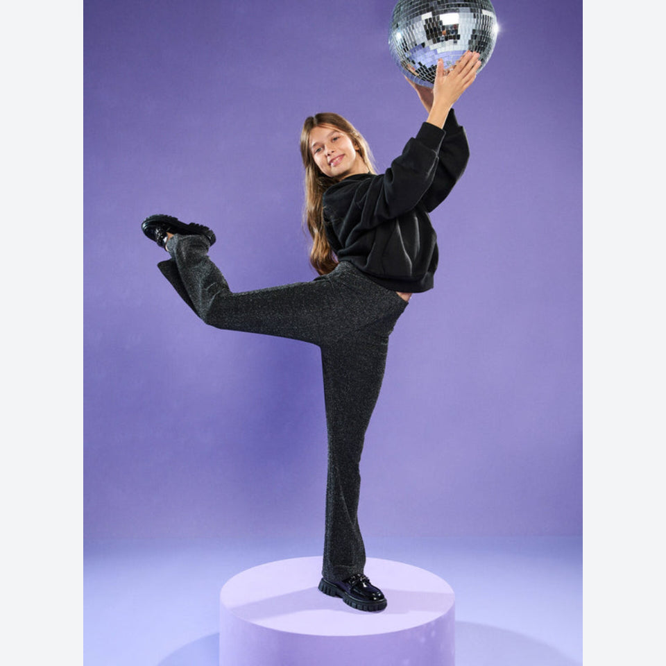A woman poses dynamically on a light purple pedestal, wearing a black sweatshirt and dark charcoal flared pants, holding a silver disco ball against a solid purple studio background.