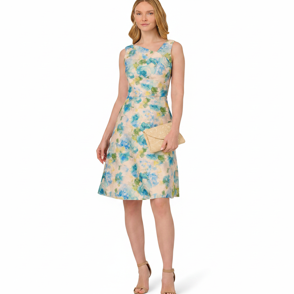 A woman modeling a sleeveless knee-length A-line dress with a blue and green watercolor floral print on a cream background, holding a beige clutch and wearing beige heels against a white studio background.