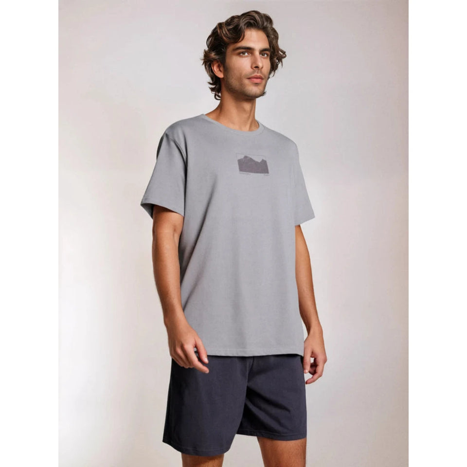 A man wearing a light gray oversized crewneck t-shirt with a small dark gray graphic on the left chest, paired with dark navy shorts, standing against a light gray studio background.