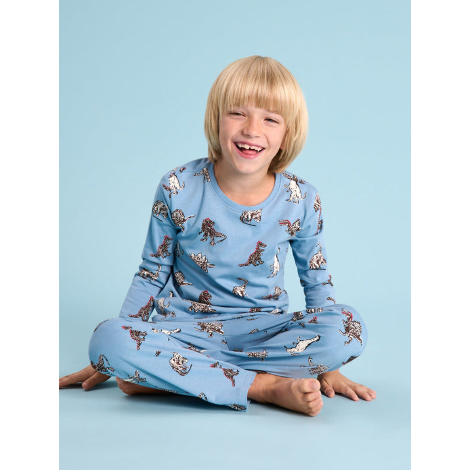 A young child with blonde hair sits cross-legged on a light blue studio background, wearing a light blue long-sleeve pajama set with a dinosaur print.