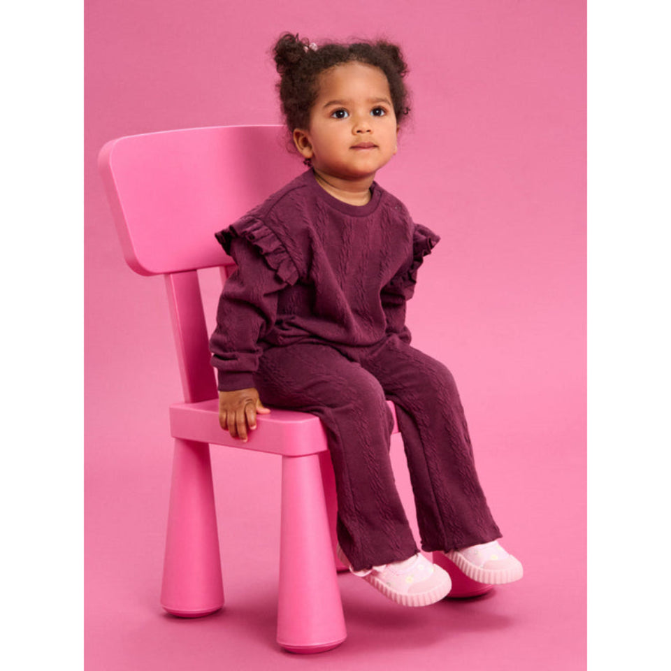 A toddler girl wearing a matching textured burgundy corduroy two-piece outfit with ruffle sleeves, sitting on a bright pink plastic chair against a solid pink background.