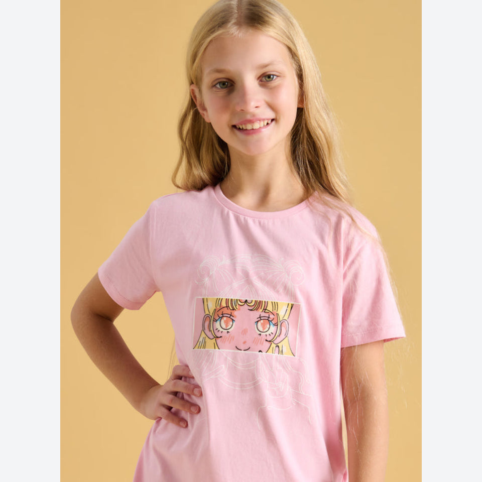 A young girl with blonde hair smiling while wearing a light pink short-sleeve t-shirt with a cartoon graphic of a girl's face on the front, set against a solid warm beige background.