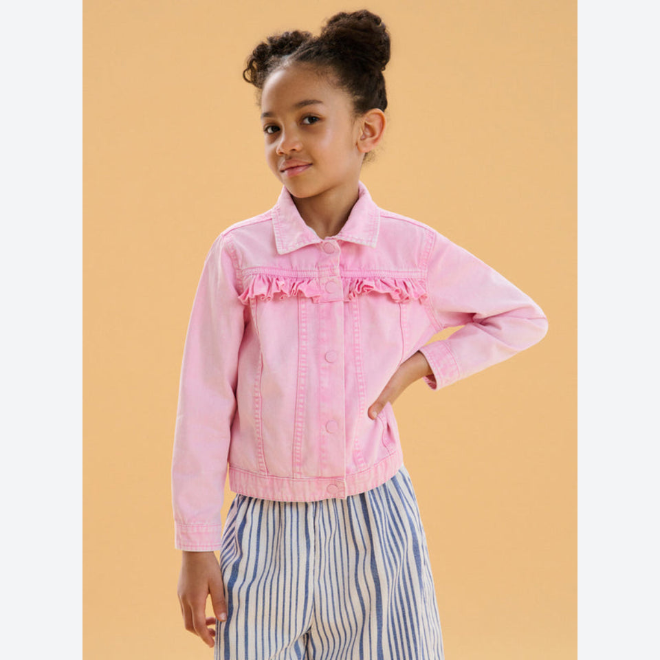 A young girl with her hair in a high bun wears a light pink denim jacket with ruffle details on the chest, paired with blue and white striped pants, set against a solid warm beige background.