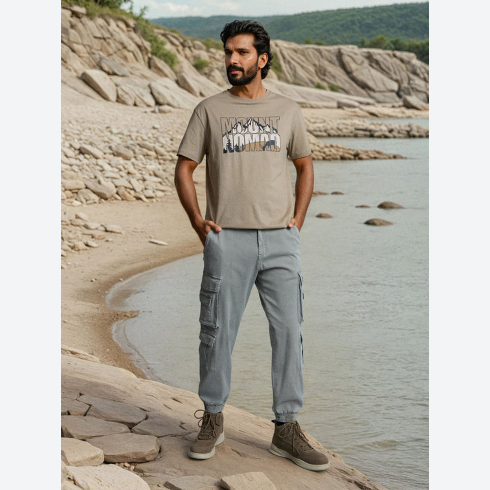 A man stands on a rocky beach shoreline wearing a beige graphic t-shirt, light gray cargo jogger pants, and brown sneakers, with water and rocky cliffs in the background.