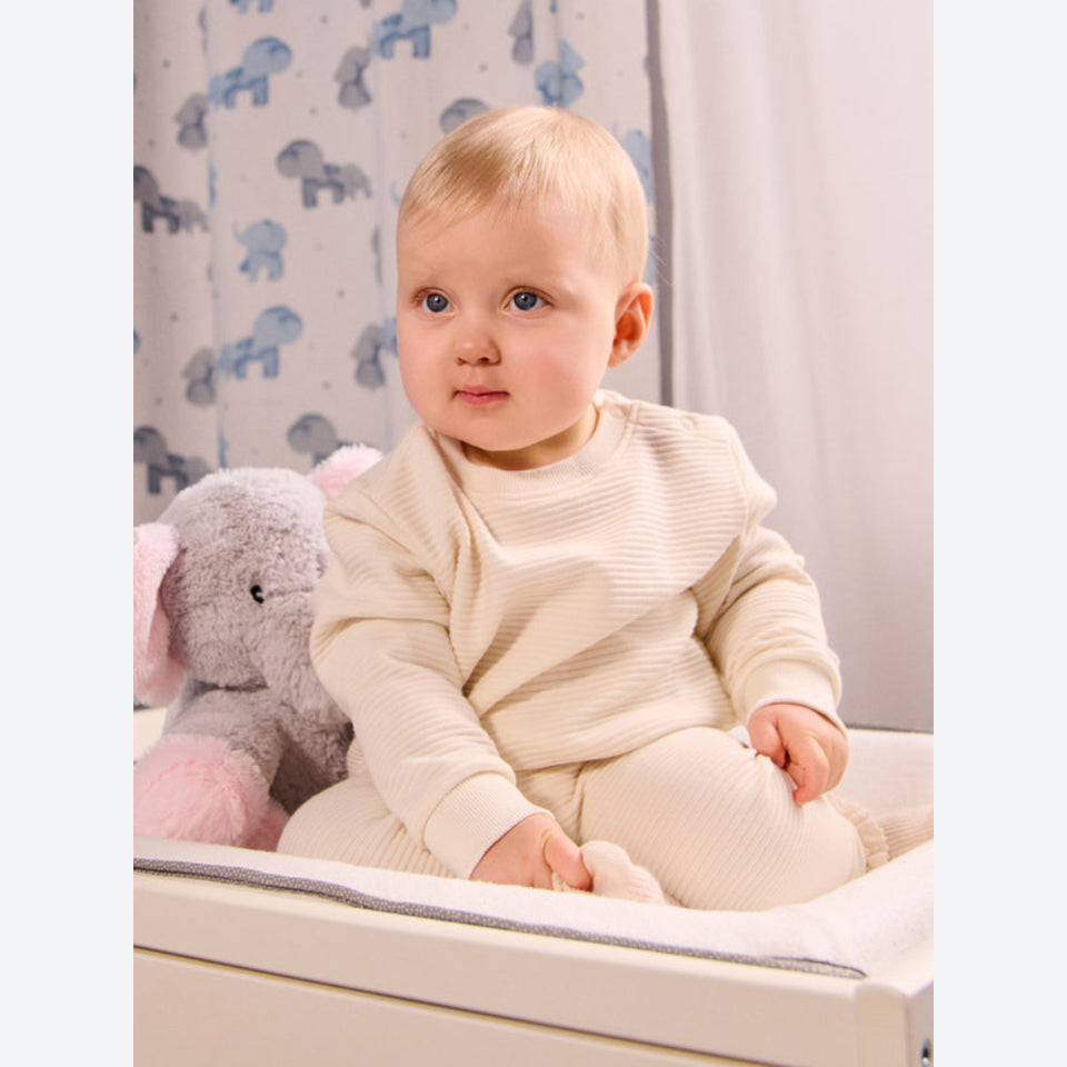 A baby sitting on a changing table wearing a cream ribbed long-sleeve top and matching cream pants, with a gray stuffed elephant toy beside them and a patterned curtain in the background.
