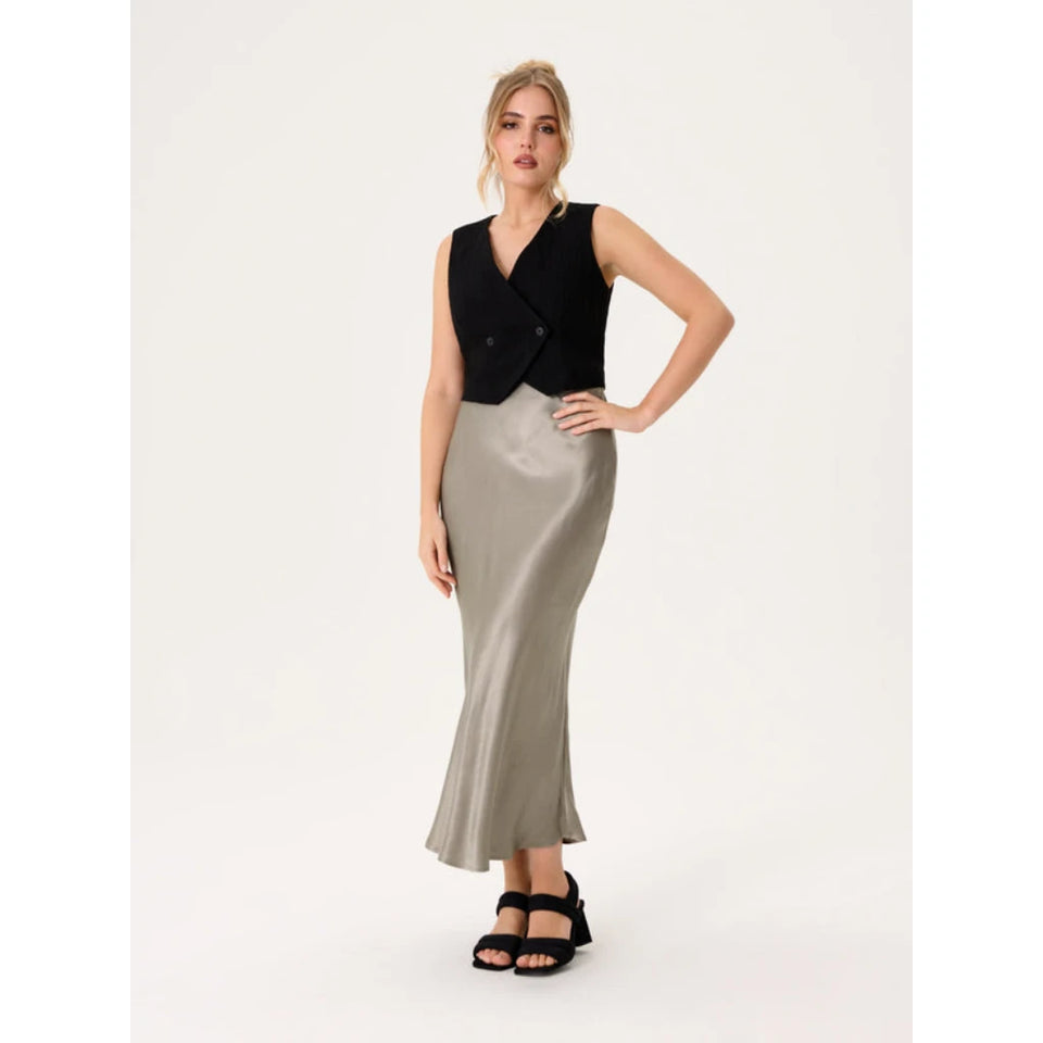 A woman modeling a sleeveless black wrap-style vest paired with a floor-length metallic silver skirt and black strappy sandals, photographed against a plain light studio background.
