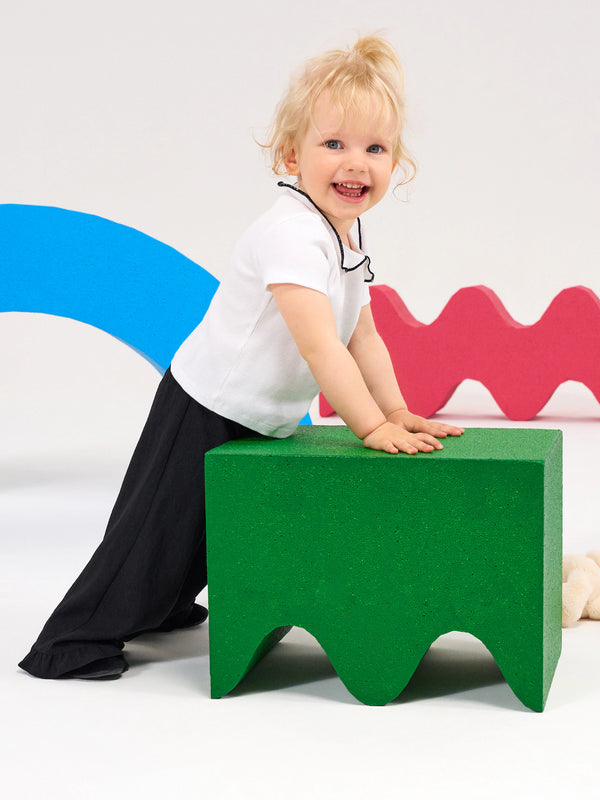 A toddler with blonde hair, wearing a white t-shirt and black pants, smiling while leaning on a green foam block in a playful studio setting.