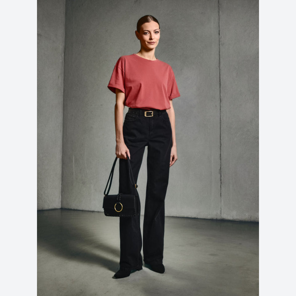 A model stands in a minimalist concrete room wearing a dusty rose short-sleeve top, black high-waisted wide-leg trousers with a gold buckle belt, and holding a black handbag.