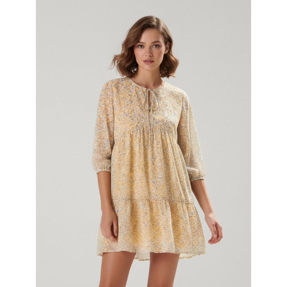 A woman modeling a light yellow tiered mini dress with a delicate blue and white floral pattern, three-quarter sleeves, and a tie-neck detail, set against a plain light gray studio background.