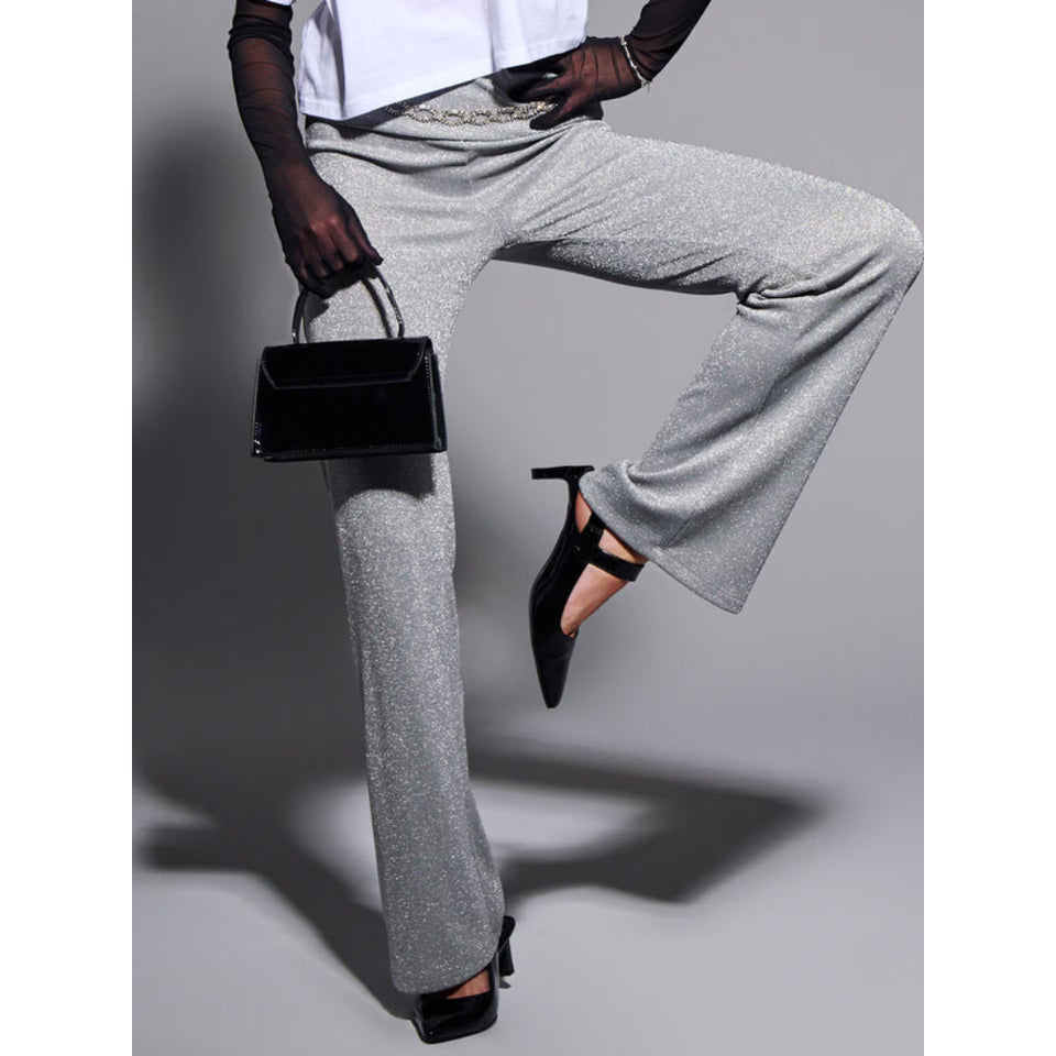 A model wearing high-waisted, wide-leg silver-gray trousers with a subtle glitter finish, styled with a white top, sheer black gloves, black pointed-toe heels, and holding a small black handbag against a gray studio background.